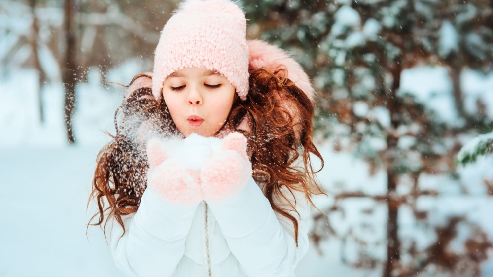 A young girl wears a white coat, a pink beanie, and pink mittens while holding snow and blowing it into the air.