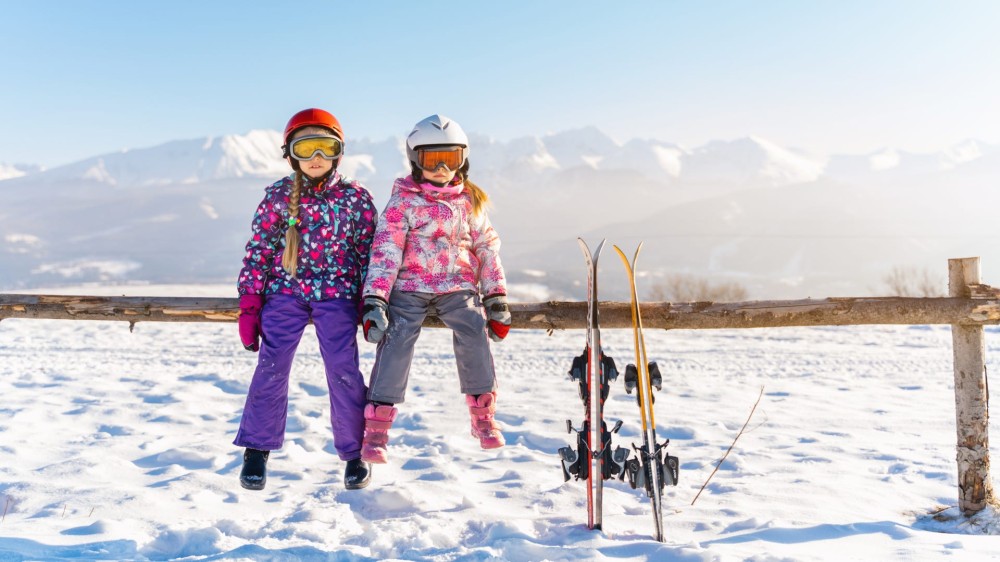 Little girls in outwear and masks with helmet sitting on wood fence with skis near against mountains